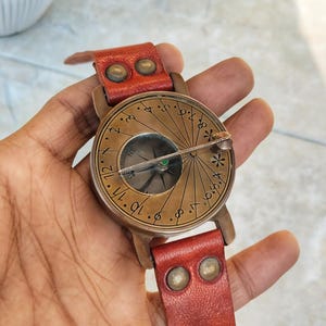 May include: A vintage-style compass watch with a bronze-colored face and a red leather band. The watch face features etched markings and a compass needle. The leather band is secured with brass-colored rivets.