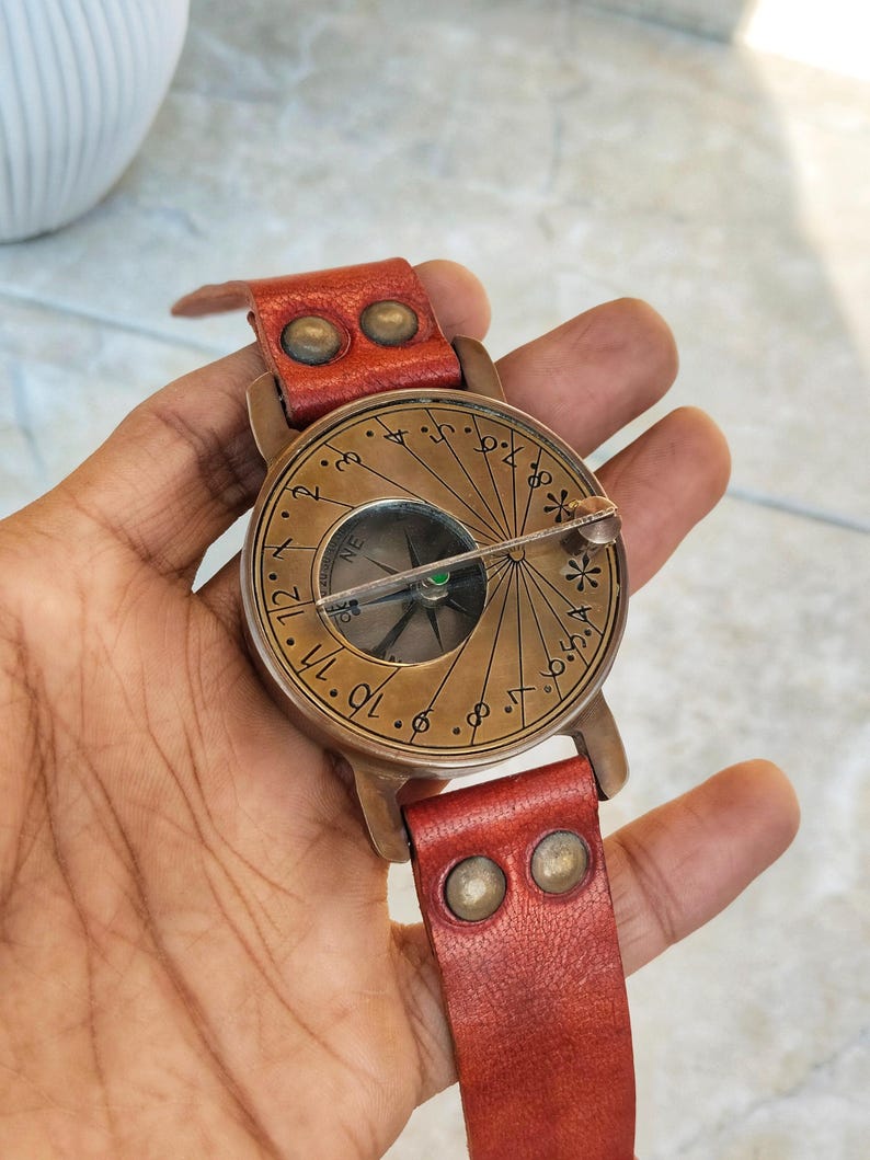 May include: A vintage-style compass watch with a bronze-colored face and a red leather band. The watch face features etched markings and a compass needle. The leather band is secured with brass-colored rivets.
