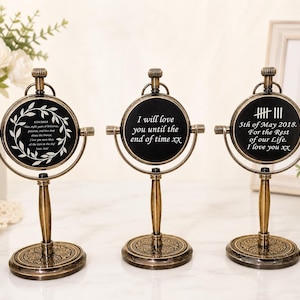 May include: Three antique-style, bronze-coloured decorative stands with round, black faces. Each stand has a different inscription in white text. The stands are on a white surface, with a vase of white flowers in the background.