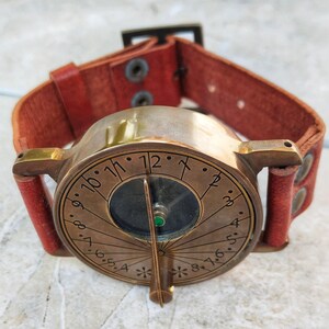 May include: A vintage-style brass sundial watch with a brown leather band. The watch face features etched numerals and a central compass. The red leather band is secured with rivets and a buckle. A unique, steampunk-inspired accessory.