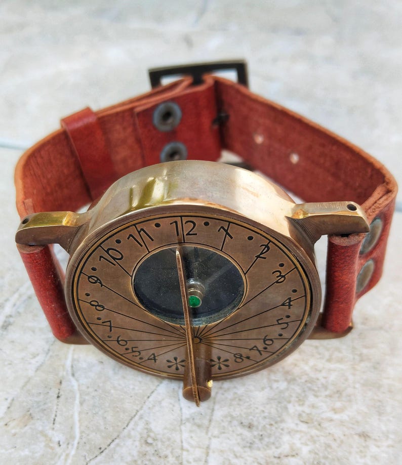 May include: A vintage-style brass sundial watch with a brown leather band. The watch face features etched numerals and a central compass. The red leather band is secured with rivets and a buckle. A unique, steampunk-inspired accessory.