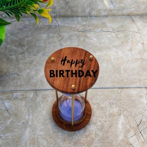 Może przedstawiać: Drewniana klepsydra z wygrawerowanym napisem "Happy Birthday" na górze. Klepsydra ma przezroczysty szklany środek wypełniony fioletowym piaskiem i jest utrzymywana przez złote metalowe podpory. Podstawa i góra wykonane są z ciemnego drewna.