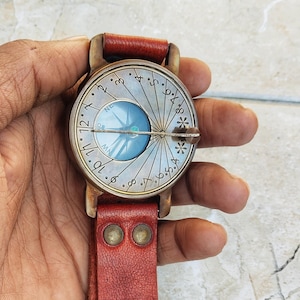 May include: A vintage-style compass watch with a round, silver-colored face featuring a blue compass rose. The watch has a red leather strap and a bronze-colored metal casing. The face has numbers and markings.