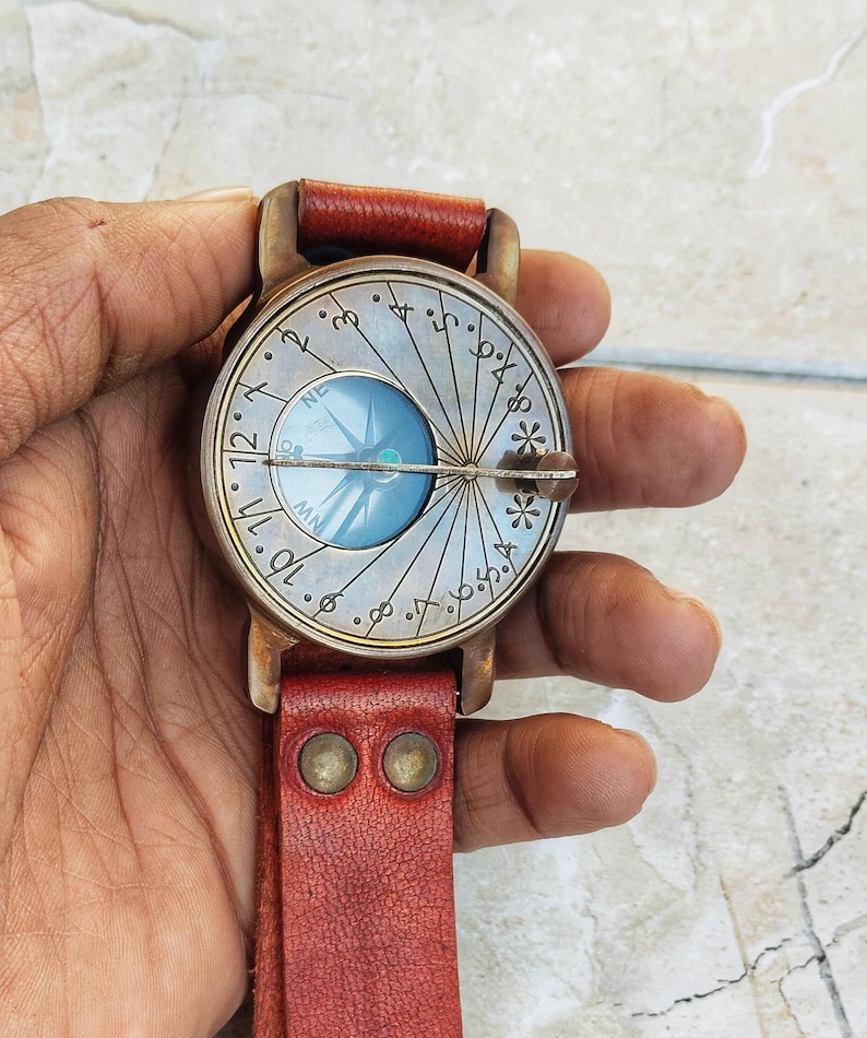 May include: A vintage-style compass watch with a round, silver-colored face featuring a blue compass rose. The watch has a red leather strap and a bronze-colored metal casing. The face has numbers and markings.