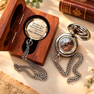 May include: Two silver pocket compasses with metal chains. One compass is inside a wooden box with the engraved text: Year After Year, You Show Me What True Love Is. Happy Anniversary. The other compass is open, showing a detailed dial face.