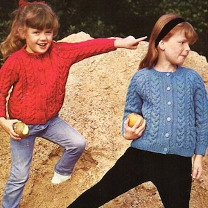 May include: Two young girls wearing knitted cardigans. The girl on the left is wearing a red cardigan and the girl on the right is wearing a blue cardigan. Both girls are holding apples.