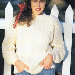 May include: A white knitted sweater with a cable knit pattern and white pom-poms. The sweater has a round neckline and long sleeves. The model is wearing blue jeans and a red scarf.