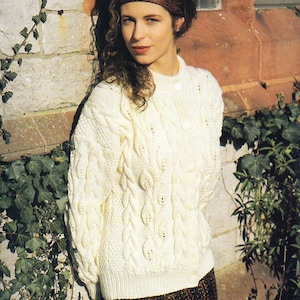 May include: A woman wearing a white cable knit sweater with a brown and black patterned skirt. She is wearing a red and brown patterned headband.