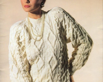 May include: A white knitted sweater with a cable knit pattern and pearl necklace. The sweater has a round neckline and long sleeves.