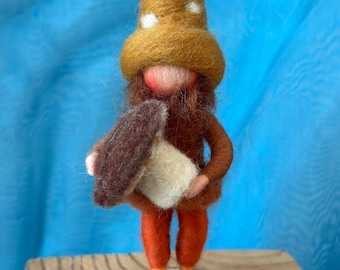 May include: A handmade felt gnome figurine with a brown body, orange pants, and a mustard-colored hat with white polka dots. The gnome holds a brown and cream-colored mushroom. The figurine is displayed on a wooden slice.