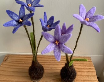 以下が含まれることがあります： 紫と青の色合いの手作りのフェルトフラワー。花は6枚の花びらと黄色の中心を持っています。茎は茶色で緑の葉が付いています。花は小さな濃い茶色のフェルトベースにセットされており、気まぐれな花の配置になっています。