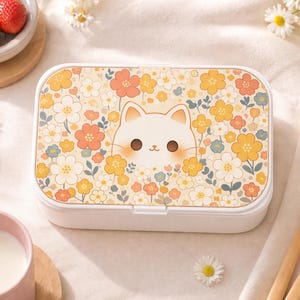 May include: A white rectangular lunchbox with a floral pattern and a cartoon cat face. The design features yellow, orange, and blue flowers on a cream background. The lunchbox is closed and sits on a white surface.