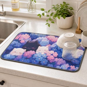 Puede incluir: Una alfombrilla rectangular para secar platos con un diseño caprichoso de gatos de dibujos animados entre flores azules y rosas. Un cuenco blanco, un vaso transparente y una taza blanca están sobre la alfombrilla.
