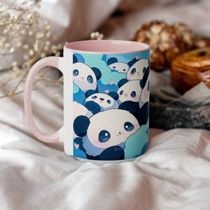 Op de afbeelding: Een witte keramische mok met een roze rand heeft een ontwerp met schattige cartoon-panda's met blauwe en witte accenten. De panda's zijn omringd door blauwe wolken en roze harten.