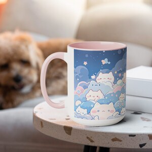 以下が含まれることがあります： ピンクの縁取りが付いた白いセラミック製のマグカップには、雲と星のある夜空に、漫画の猫が描かれた幻想的なデザインが施されています。マグカップは薄い茶色のテーブルの上に置かれています。