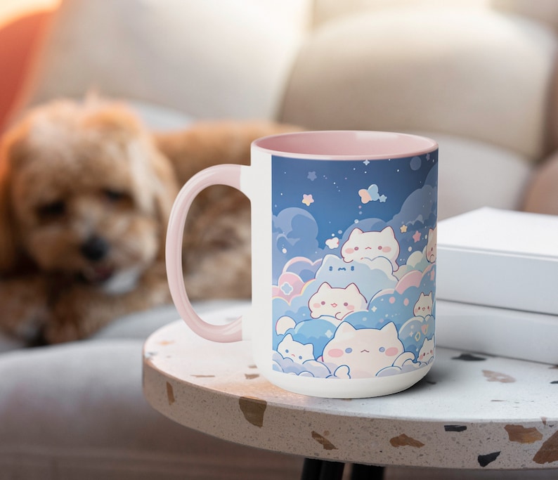 以下が含まれることがあります： ピンクの縁取りが付いた白いセラミック製のマグカップには、雲と星のある夜空に、漫画の猫が描かれた幻想的なデザインが施されています。マグカップは薄い茶色のテーブルの上に置かれています。