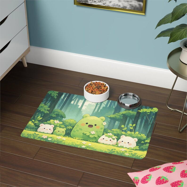 May include: A rectangular pet food mat featuring a whimsical forest scene with cartoon bears and a green, smiling creature. The mat has a white ceramic bowl filled with pet food and a stainless steel bowl. The mat is placed on a dark wood floor.