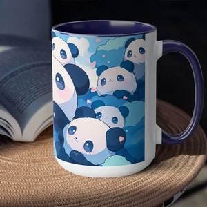 Op de afbeelding: Een witte keramische mok met een blauwe rand en handvat. De mok heeft een ontwerp met meerdere cartoonpanda's met blauwe en witte wolken en harten. De panda's lachen allemaal en hebben zwarte ogen.