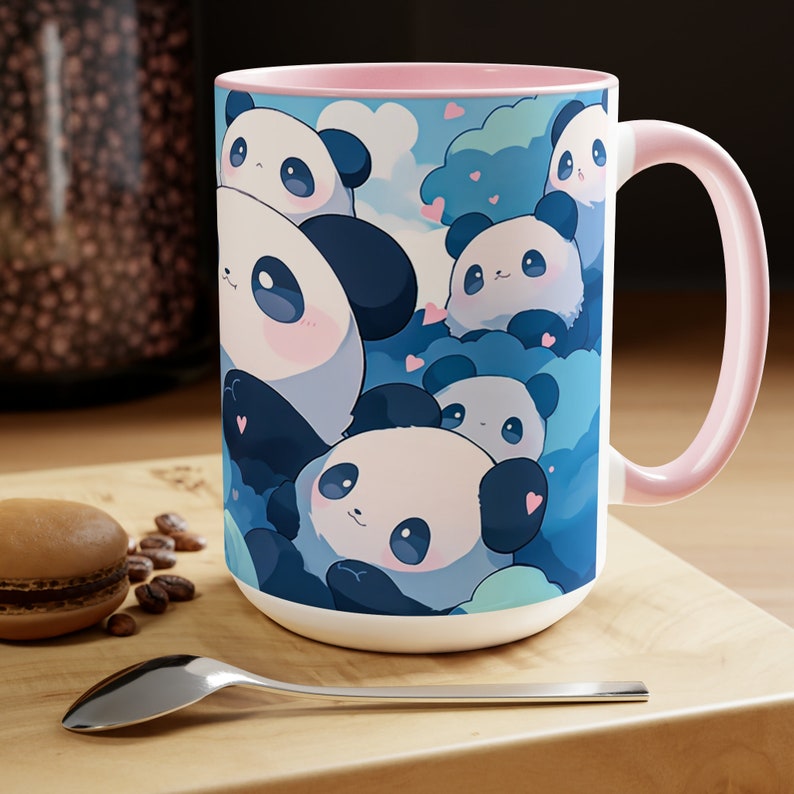 Op de afbeelding: Een witte keramische mok met een roze rand en handvat. De mok heeft een kleurrijke illustratie van meerdere cartoonpanda's met blauwe en roze accenten. De panda's zijn omringd door blauwe wolken en roze harten.