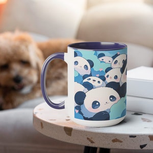 Op de afbeelding: Een witte keramische mok met een paarse rand heeft een ontwerp met meerdere schattige cartoonpanda's met blauwe en witte accenten. De panda's worden omringd door blauwe en witte wolken met roze harten.