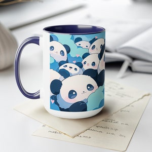 Op de afbeelding: Witte keramische mok met een blauwe rand en een ontwerp van schattige cartoon-panda's op een blauwe en witte achtergrond.