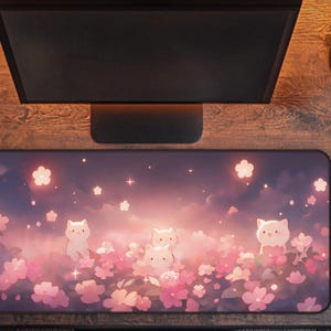 May include: A rectangular mousepad featuring a whimsical design of white cat characters amidst pink cherry blossoms and a dreamy sky. The pad is designed for computer use and is surrounded by a wooden desk, a computer monitor, and a mouse.