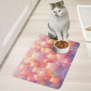 May include: A white cat sits on a pink and purple pet food mat with a white bowl of dry cat food. The mat has a pattern of cartoon bears.