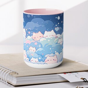 以下が含まれることがあります： ピンクの縁取りが付いた白いセラミック製のマグカップには、星が散りばめられた青とピンクの雲のような空に、漫画の猫が描かれた幻想的なデザインが施されています。