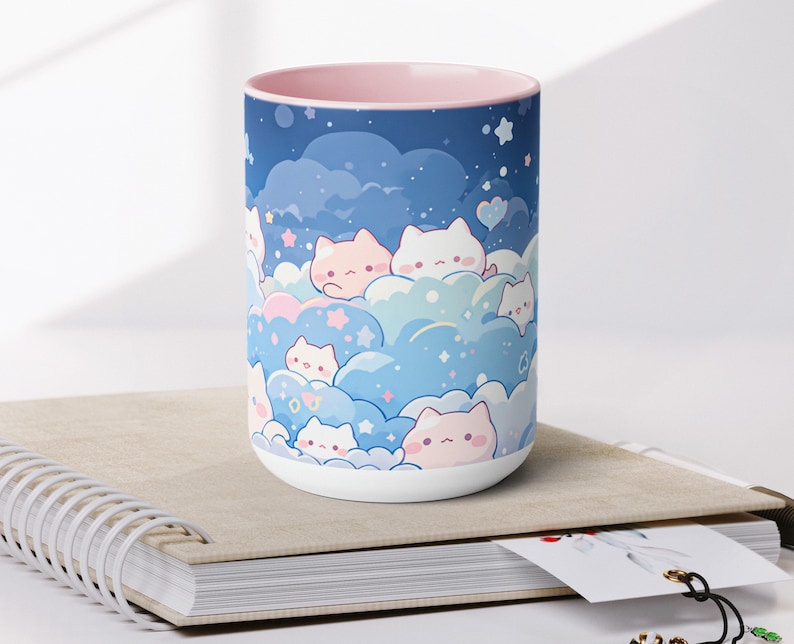 以下が含まれることがあります： ピンクの縁取りが付いた白いセラミック製のマグカップには、星が散りばめられた青とピンクの雲のような空に、漫画の猫が描かれた幻想的なデザインが施されています。
