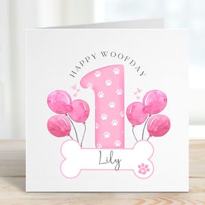 May include: Pink birthday card with a paw print design. The card says "Happy Woofday" and features a pink number one with paw prints. The card is personalised with the name "Lily".