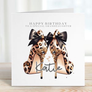 May include: A birthday card featuring a pair of leopard print high heels with black bows and gold detailing. The card is inscribed with "Happy Birthday to a Special Granddaughter" and the name "Katie".