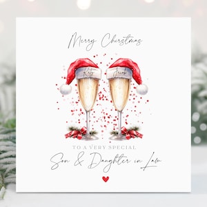 May include: A white Christmas card with a red and white Santa hat on top of two champagne glasses. The text on the card reads "Merry Christmas to a very special Son & Daughter in Law".