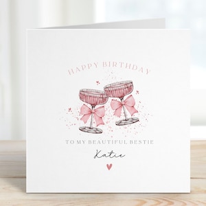 May include: A white birthday card with pink text reading "HAPPY BIRTHDAY" and "TO MY BEAUTIFUL BESTIE Katie". It features two pink champagne glasses with bows. A pink heart is at the bottom.