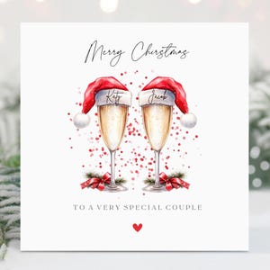 May include: A Christmas card with a watercolor illustration of two champagne flutes topped with Santa hats. The flutes are surrounded by red and pink confetti. The text on the card reads "Merry Christmas" and "To a very special couple".