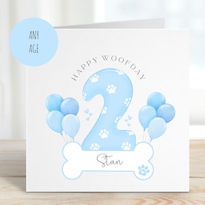 May include: A blue and white birthday card with a paw print design. The card says "Happy Woofday" and features a blue number 2 with white paw prints. The card is personalised with the name "Stan".