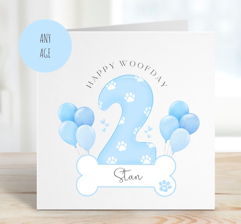 May include: A blue and white birthday card with a paw print design. The card says "Happy Woofday" and features a blue number 2 with white paw prints. The card is personalised with the name "Stan".