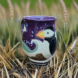 May include: A handmade ceramic mug with a purple exterior and a whimsical puffin design. The mug features a black, white, and orange puffin, with a wave and starry sky design. The mug is set against a backdrop of tall grass.