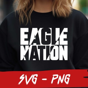 May include: A black sweatshirt with white text that reads "EAGLE NATION" in a bold, graphic font. The text is layered and appears to be cut out of the fabric. The sweatshirt is being worn by a person, but the person's face is not visible. The image is set against a blurred background of green foliage.