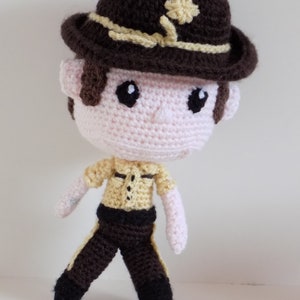 May include: A crocheted plush toy of a person wearing a brown hat with a yellow band and a yellow flower, a yellow shirt, and brown pants.