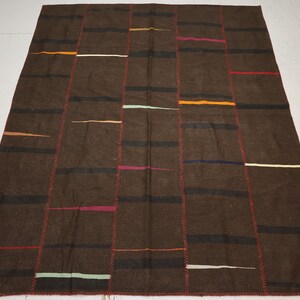 Puede incluir: Una alfombra marrón con un diseño de patchwork. La alfombra presenta rayas verticales de varios colores, incluyendo negro, naranja, rojo y blanco. Los bordes están cosidos con hilo rojo y la alfombra tiene forma rectangular.