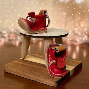 May include: Two red leather boot keychains on a wooden stand. The boots are detailed with stitching and have a metal ring for attaching keys.