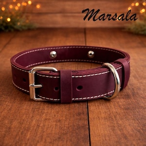 Può includere: Un collare per cani in pelle bordeaux con cuciture bianche e finiture color argento. Il collare è dotato di una chiusura a fibbia e di un anello a D per l'attacco del guinzaglio. La parola "Marsala" è scritta in corsivo.