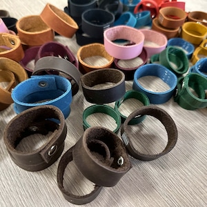 May include: Assortment of leather bracelets in various colours, including brown, blue, green, pink, and black. The bracelets are stacked and arranged on a wooden surface.