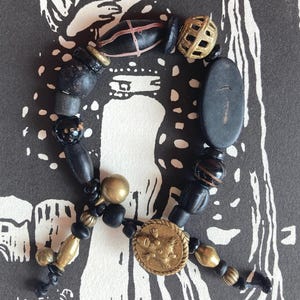 Könnte beinhalten: Ein schwarzes und goldenes Perlenarmband mit verschiedenen Perlen, darunter eine große schwarze ovale Perle, eine kleinere schwarze Perle mit goldener Akzentuierung und eine goldene Perle mit geometrischem Muster. Das Armband befindet sich auf einem schwarz-weißen Hintergrund.