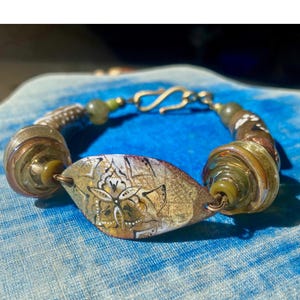 May include: A handcrafted bracelet featuring a central, leaf-shaped bead with a floral design. The bracelet incorporates spiral-shaped glass beads in shades of brown and green, connected by a silver chain and clasp.