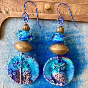 Puede incluir: Un par de pendientes colgantes con dijes circulares azules y morados, uno con diseño floral y otro con motivo de búho. Cada pendiente incluye una cuenta de tono latón, una cuenta de vidrio azul y un gancho de metal azul junto a una regla.