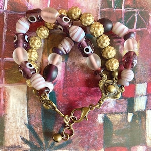 Peut inclure: Un bracelet à double rang de perles violettes, blanches et dorées. Le bracelet est doté d'un fermoir en laiton et d'un charme en forme de cloche.