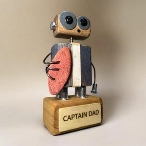 Op de afbeelding: Een houten robotsculptuur met een blokvormig lichaam en hoofd. De robot heeft grote, ronde ogen van metalen ringen en een rood schild. De basis van de robot leest "CAPTAIN DAD".