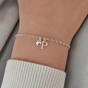 Puede incluir: Una pulsera de cadena plateada con dos dijes colgantes: un corazón pequeño, liso y pulido, y una letra P mayúscula. La pulsera está formada por eslabones redondos y delicados, y se asegura alrededor de la muñeca con un cierre sencillo.
