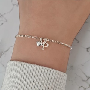 Könnte beinhalten: Ein silbernes Armband mit filigraner Kette. Es hat einen Herzanhänger und einen Buchstabenanhänger "P". Das Armband wird am Handgelenk getragen, mit einem weißen Pullover im Hintergrund.